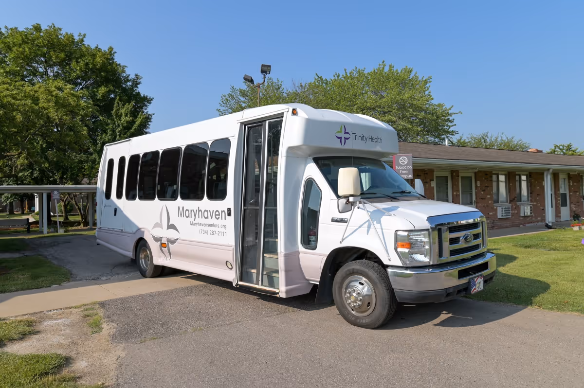 A white shuttle bus parked outside a single-story brick building with a green lawn and trees in the background. The bus has the name Maryhaven and contact information on its side, along with the Trinity Health logo on the front. The bus door is open.