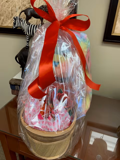 A gift basket wrapped in clear plastic with a large red bow on top, placed on a wooden table. Behind the basket, there is a decorative zebra sculpture and framed artwork on the wall.