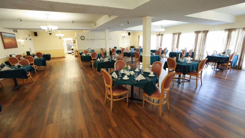 Spacious dining room with multiple round tables covered with dark green tablecloths, each set with white napkins, cups, and small flower arrangements. The room has wooden flooring, large windows with sheer curtains allowing natural light, and beige walls with a clock and wall decor.