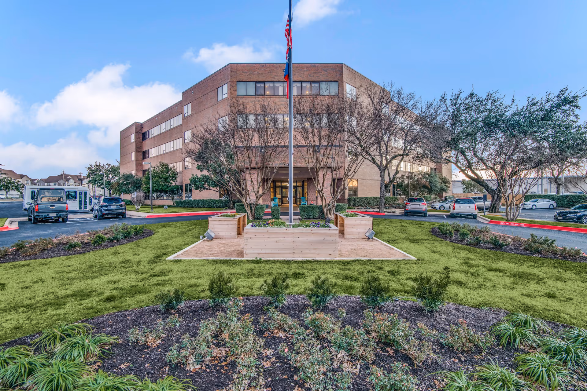 Exterior view of a multi-story brick building with a flagpole and landscaped garden in front. Several cars are parked along the driveway surrounding the garden area under a partly cloudy sky.