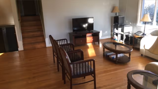 A living room with wooden flooring featuring two wooden armchairs with cushions, a glass-top coffee table, a TV on a wooden stand, a small shelving unit with a lamp, and a window letting in natural light. There is also a staircase leading up in the background.