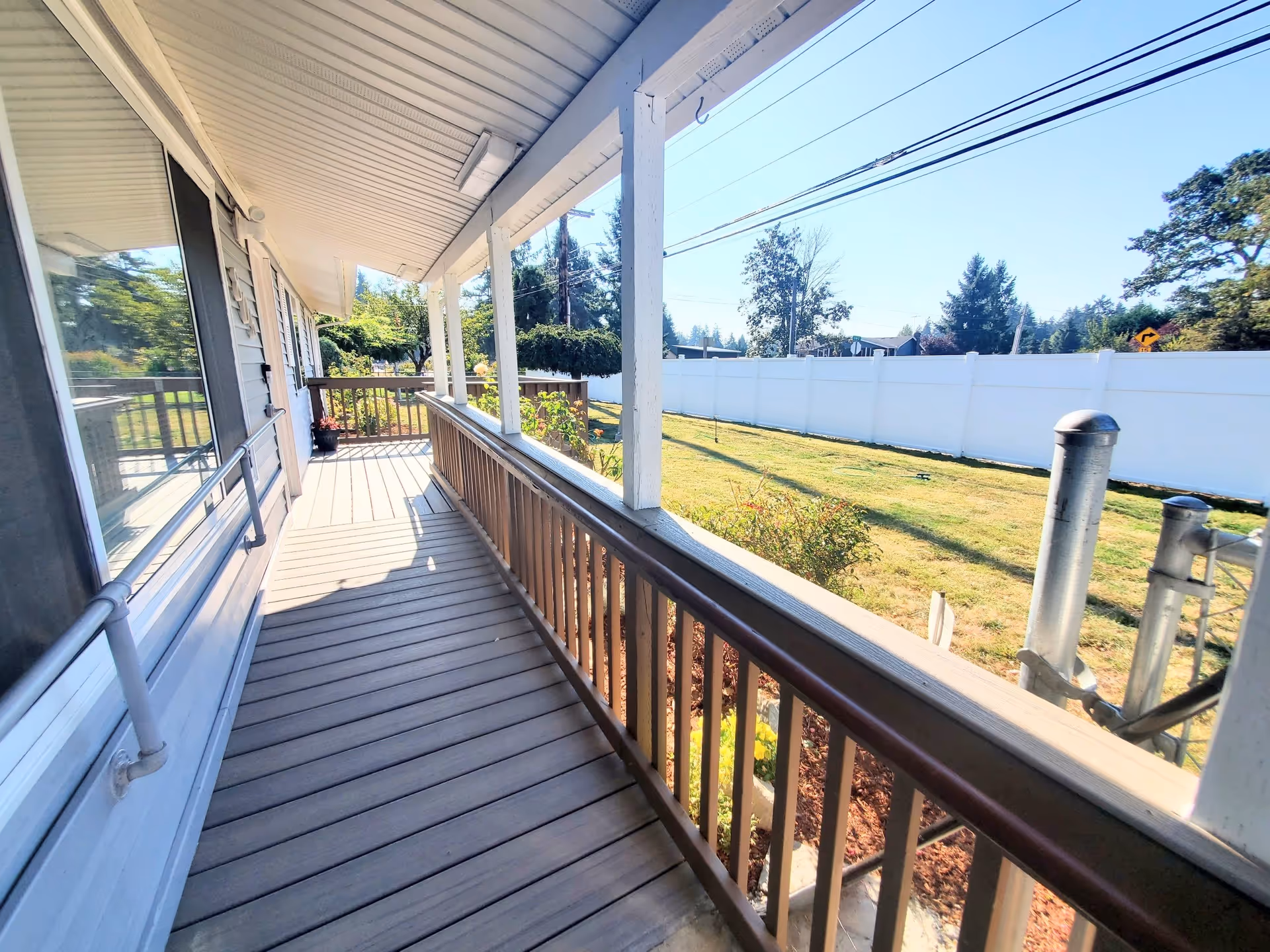 Covered wooden porch with railings and a handrail along the side of a building, overlooking a grassy yard with a white fence and trees in the background under a clear blue sky.