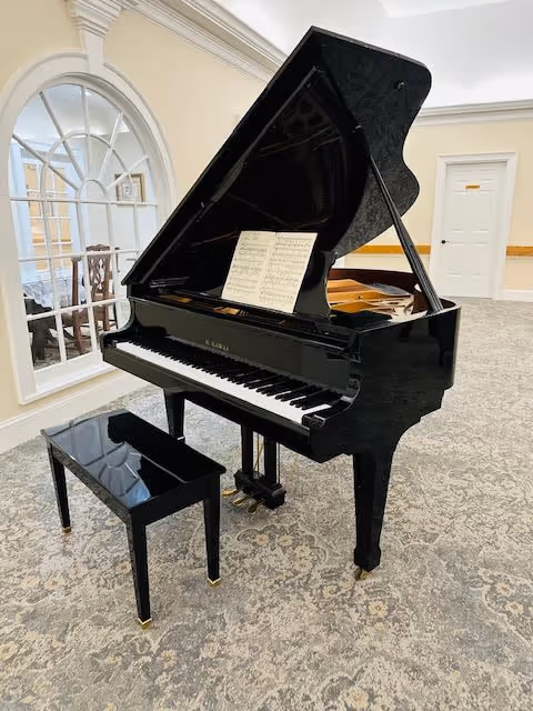 A black grand piano with its lid open and a sheet of music on the stand, positioned on a patterned carpet in a well-lit room with cream-colored walls. A matching black piano bench is in front of the piano. In the background, there is a large arched window and a white door with a wooden handrail along the wall.