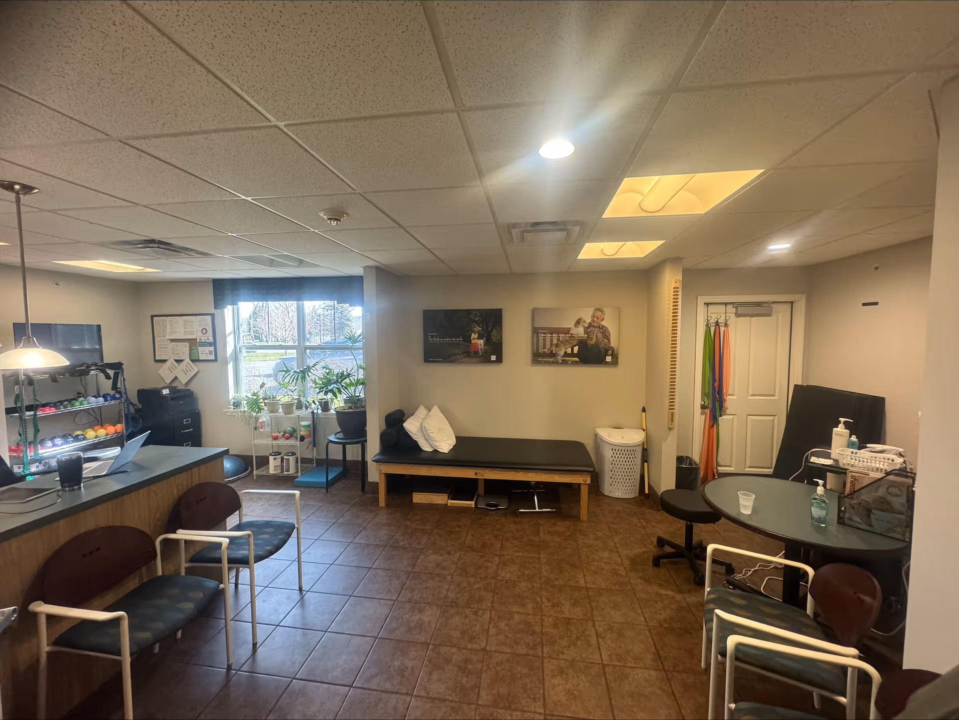 Interior common room with a reception desk, seating, exercise equipment, a treatment bench, and potted plants.