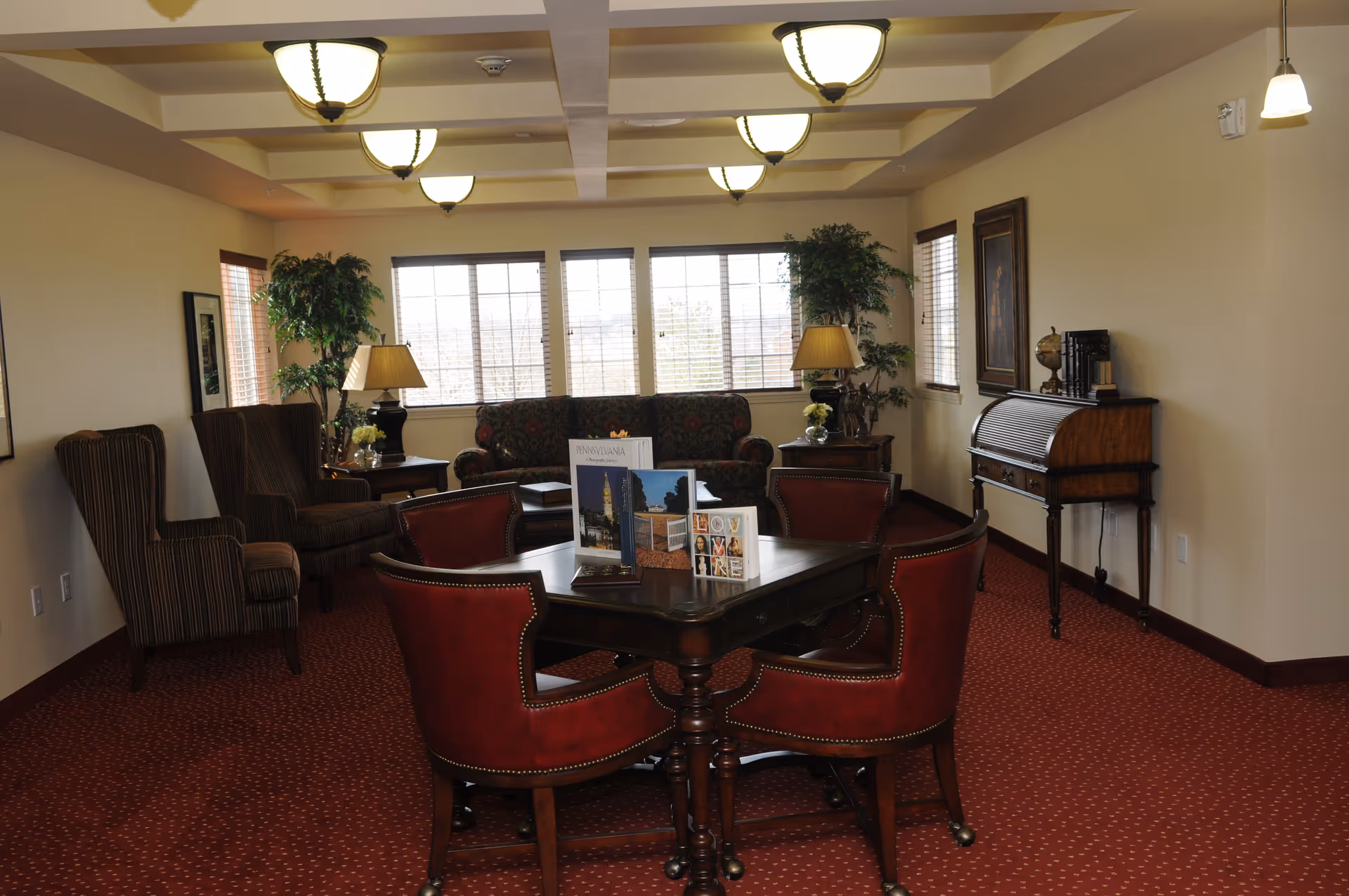 A cozy living room area with a dark wooden table surrounded by four red leather chairs in the foreground. Behind the table, there are two striped armchairs and a floral patterned sofa near large windows letting in natural light. The room has beige walls, red carpet with a subtle pattern, two table lamps on side tables, potted plants, framed artwork on the walls, and a wooden roll-top desk against the right wall.