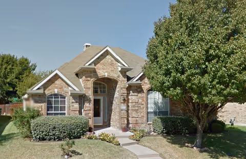Front exterior view of a single-story brick house with a gabled roof, a small front porch, and a pathway leading to the entrance. The yard has neatly trimmed bushes, a tree on the right side, and a well-maintained lawn under a clear blue sky.