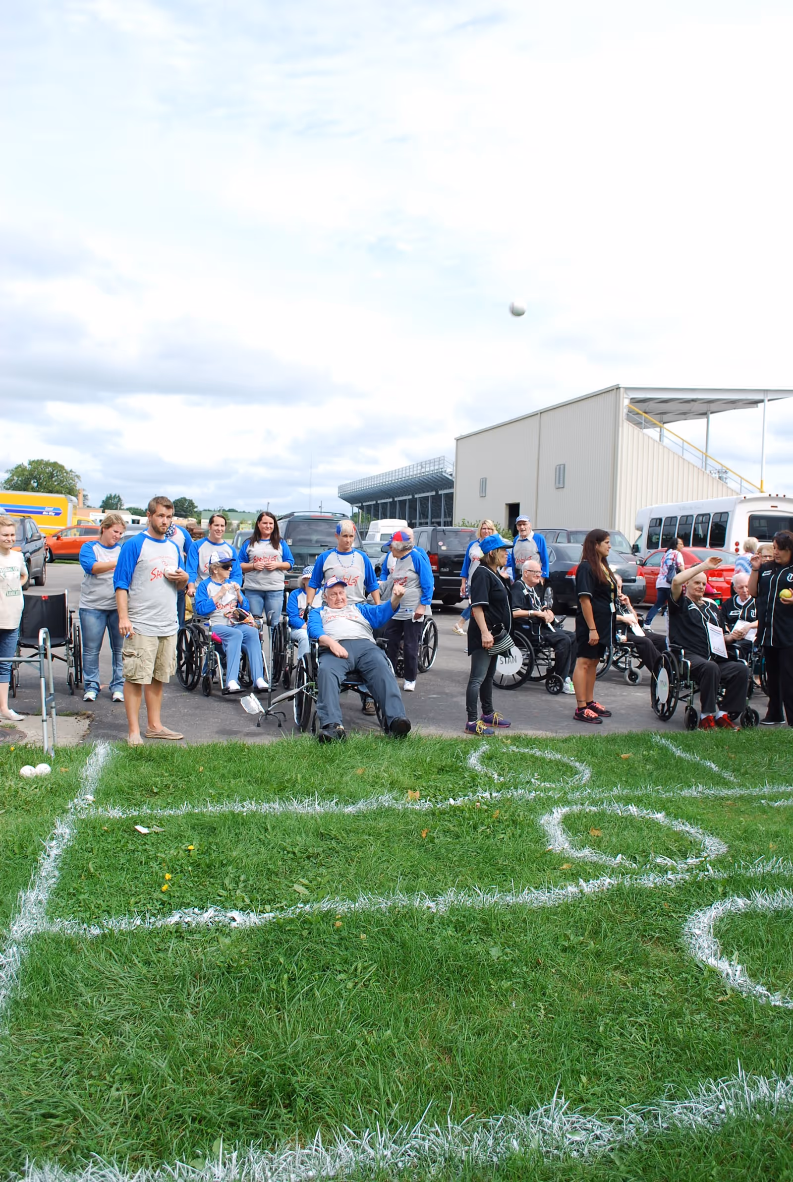 A group of people, some in wheelchairs and others standing, gathered outdoors on a grassy area with white chalk markings for a game. They appear to be participating in an organized activity or event, with a building and parked vehicles in the background under a cloudy sky.