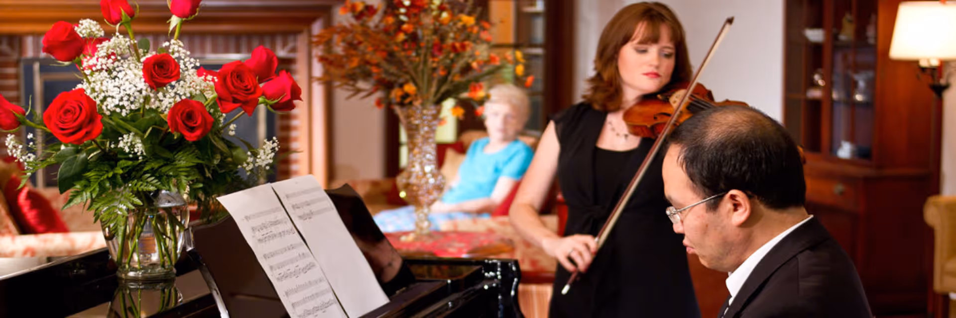 A pianist and a violinist perform in a cozy lounge area with a vase of red roses and a seated older woman in the background.