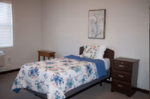 A simple bedroom with a single bed dressed in floral bedding, two wooden nightstands, a framed picture on the wall, and a window.