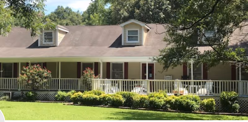 A single-story residential building with a large covered porch featuring white railings and outdoor seating. The building has a beige exterior with dark red shutters and dormer windows on the roof. In front of the porch, there are neatly trimmed bushes and a well-maintained green lawn. Trees surround the building, providing shade.