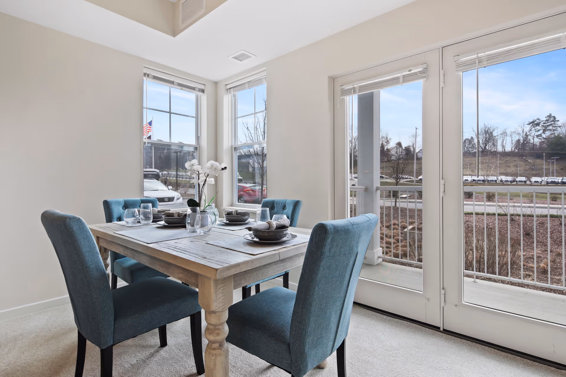 A dining area with a wooden table set for four, featuring blue upholstered chairs, glassware, and a small floral centerpiece. The room has large windows and glass doors that open to an outdoor view with a railing and a parking lot visible outside.