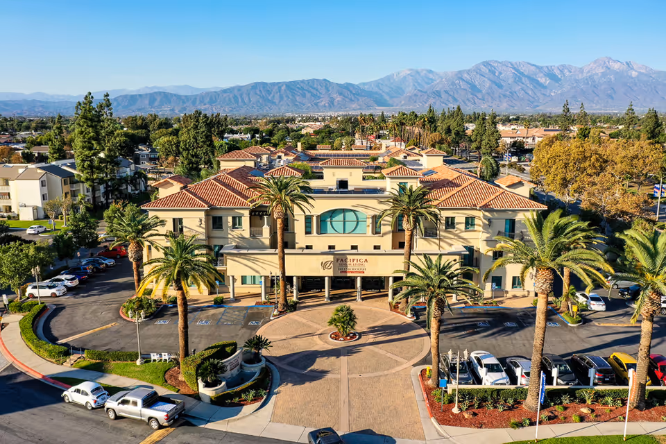 Aerial view of a senior living facility with a large beige building featuring red-tiled roofs, surrounded by palm trees and a parking lot with several cars. Mountains are visible in the background under a clear blue sky.
