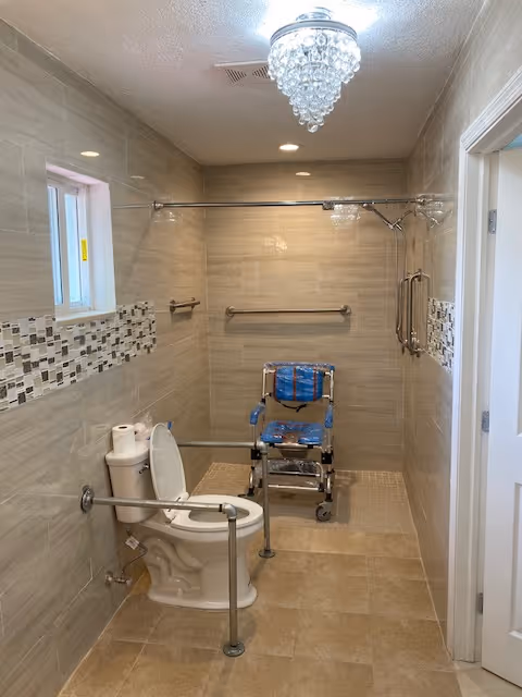 Accessible bathroom with beige tiled walls and floor, featuring a toilet with support rails, a shower area with a shower chair, grab bars, a small window, and a crystal chandelier on the ceiling.