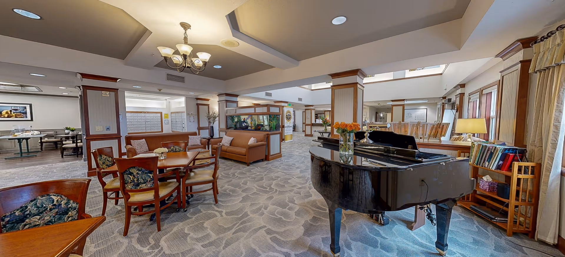 A spacious and well-lit senior living facility common area featuring a grand piano with a vase of flowers on top, several wooden tables and chairs with floral cushions, comfortable sofas, an aquarium, and bookshelves. The room has a patterned carpet, recessed lighting, and a chandelier, creating a warm and inviting atmosphere.