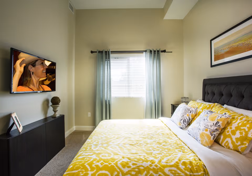 A bright bedroom with a large bed covered in yellow and white patterned bedding and multiple pillows. There is a window with light blue curtains letting in natural light. A flat-screen TV is mounted on the wall opposite the bed, showing a woman wearing a hat. A black cabinet below the TV holds a small framed photo and a decorative item. A framed landscape painting hangs above the bed's tufted dark headboard.