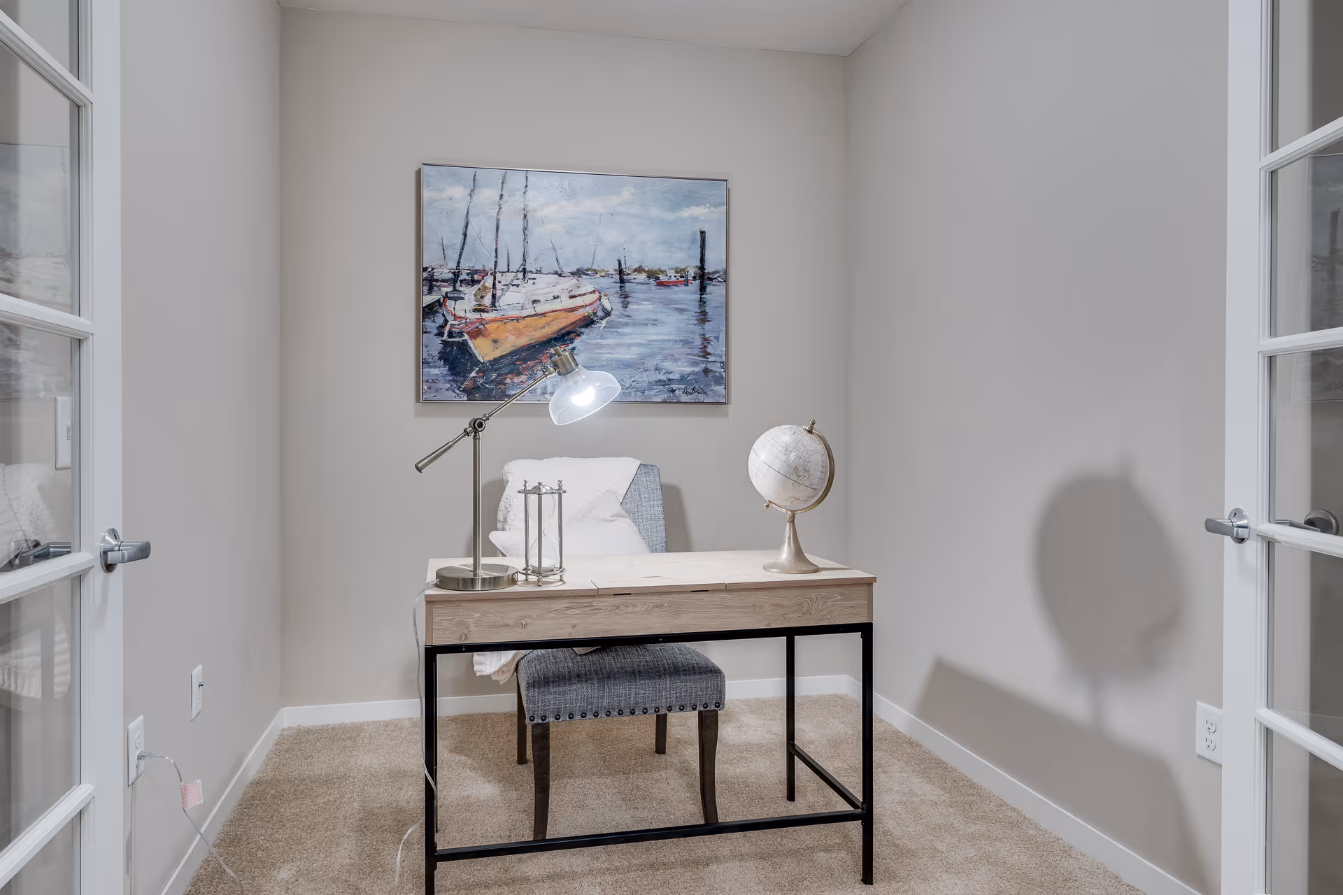 Small study with a wooden desk and chair, a lamp and globe, and a sailboat painting on the wall viewed through glass French doors.