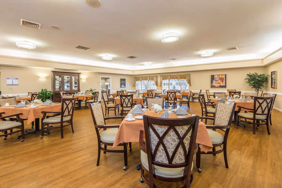 A spacious dining room with multiple round tables covered with orange tablecloths, each set with plates, cups, and folded napkins. The room has wooden flooring, cushioned chairs with patterned backs, large windows with valances, and soft ceiling lighting. There are paintings on the walls and a wooden cabinet with glass doors in the background.