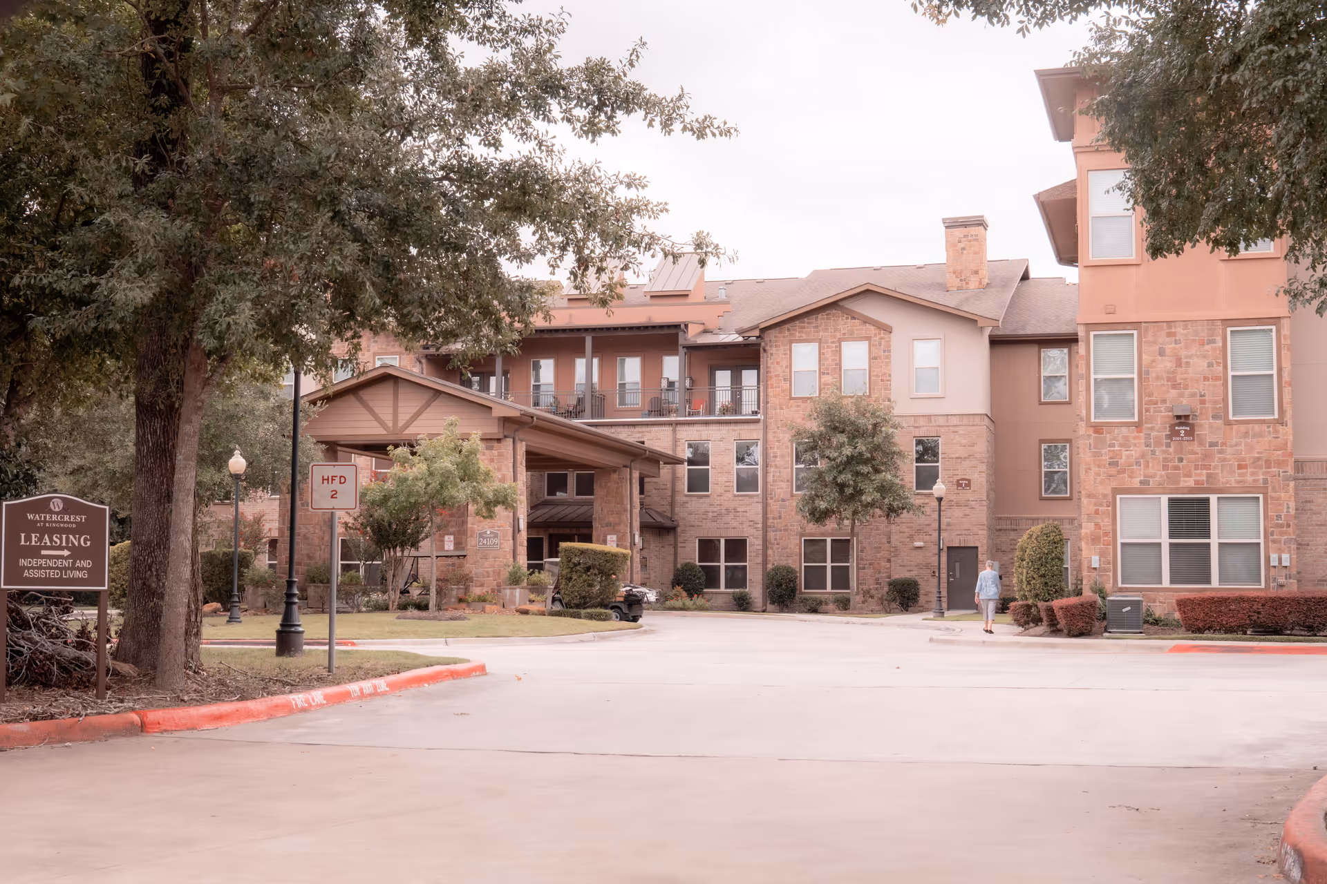 Front exterior of a multi-story brick assisted living building with a covered entrance, driveway, trees, and a person walking nearby.