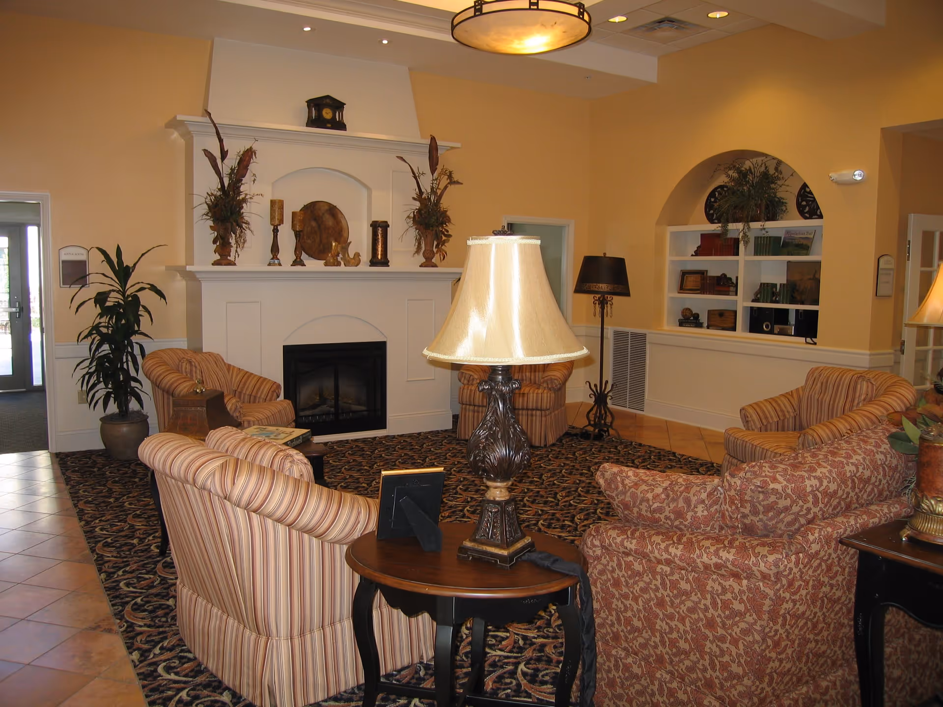 A cozy living room area with several upholstered armchairs arranged around a patterned carpet. There is a white fireplace with decorative items on the mantel, including candles and plants. A table lamp with a beige shade sits on a small wooden table in the foreground. The walls are painted a warm yellow, and there are built-in shelves with books and decorative items in an arched alcove.