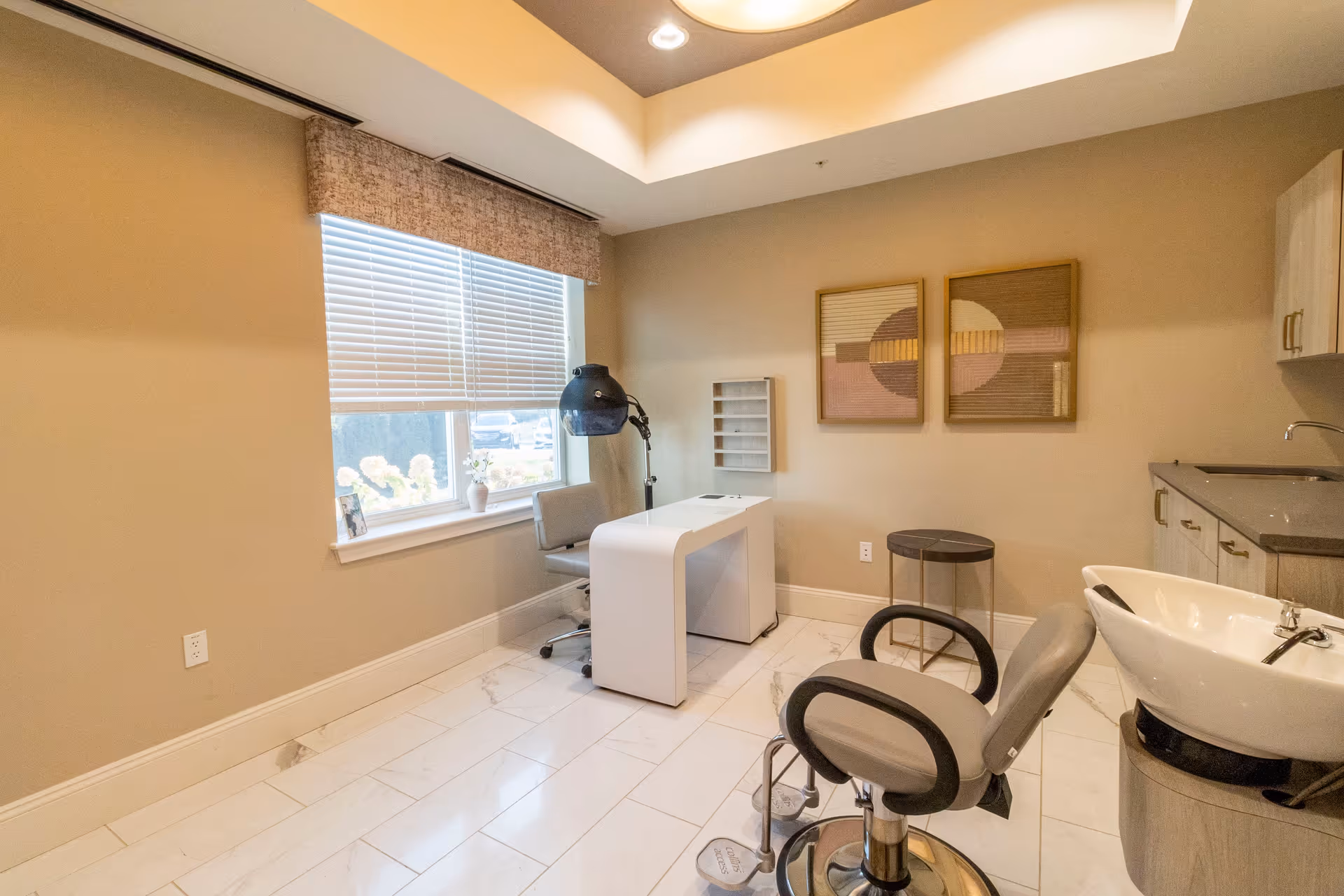 A bright and clean salon room with a large window covered by blinds, a white manicure table with a gray chair, a black hair dryer hood, a salon chair in front of a white sink, a small round side table, and two abstract framed artworks on the beige wall.
