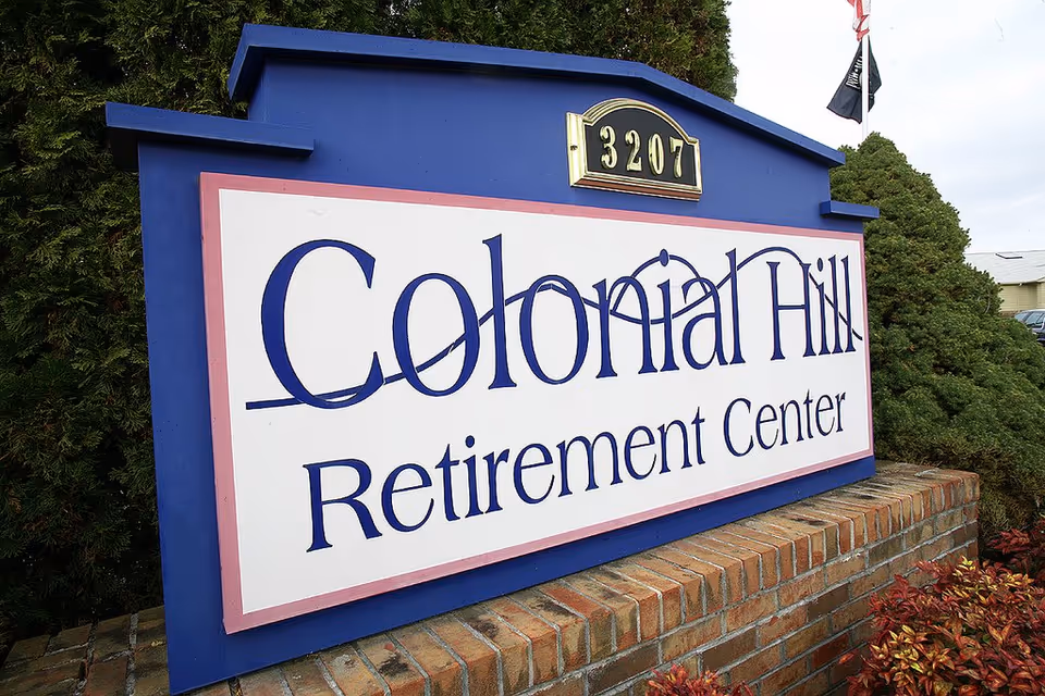 Blue-framed Colonial Hill Retirement Center sign mounted on a brick base beside evergreens