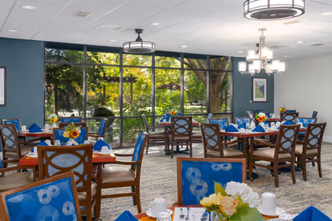 A bright dining room with multiple wooden tables and chairs featuring blue patterned cushions. Each table is set with blue napkins, white cups, and floral centerpieces. Large floor-to-ceiling windows reveal a green outdoor garden area with trees and sunlight filtering through. Modern light fixtures hang from the ceiling, and a framed picture is visible on the wall.