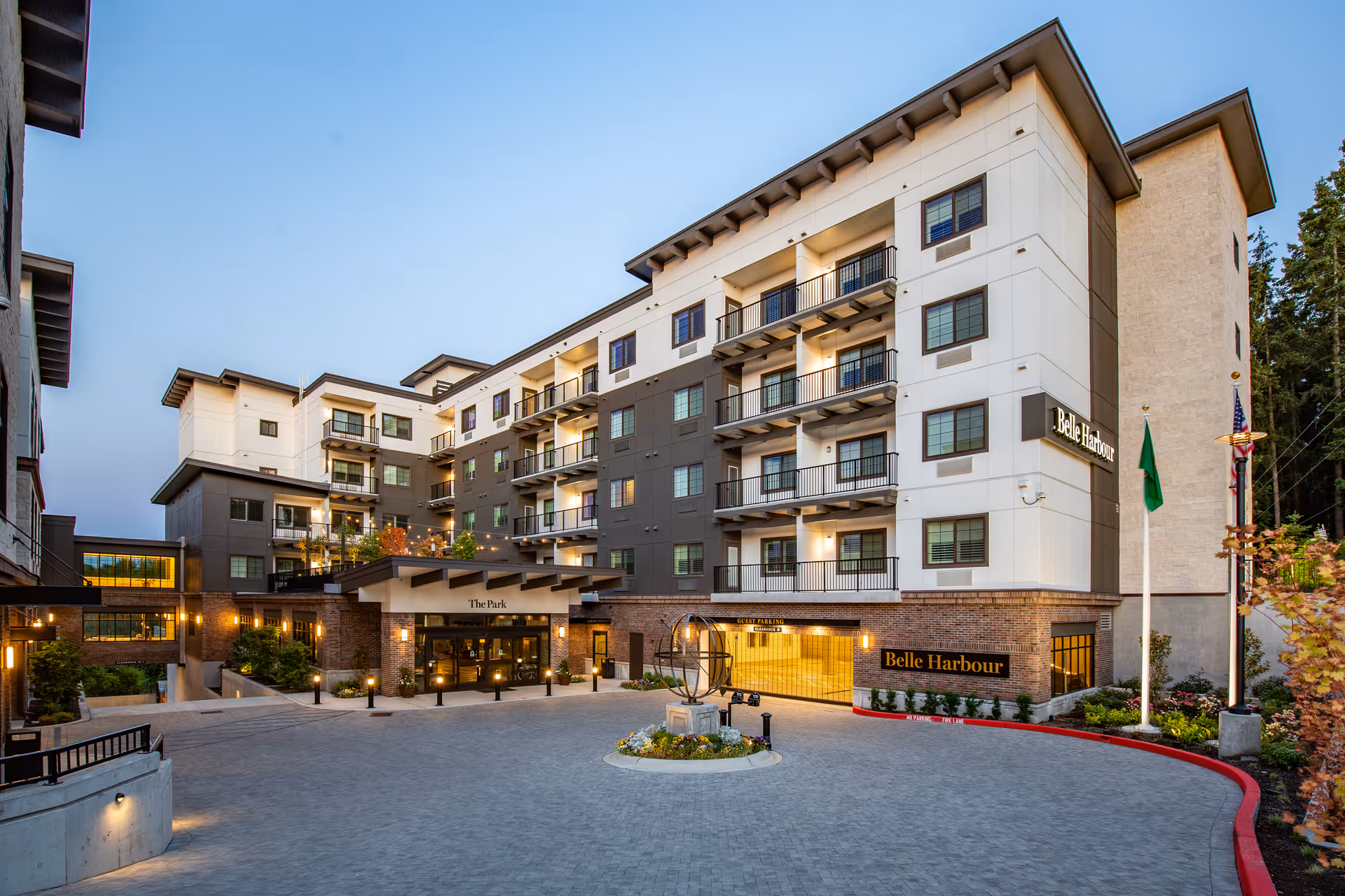 Exterior view of The Park at Belle Harbour Assisted Living Community building during early evening with well-lit entrance, balconies, and landscaped surroundings.