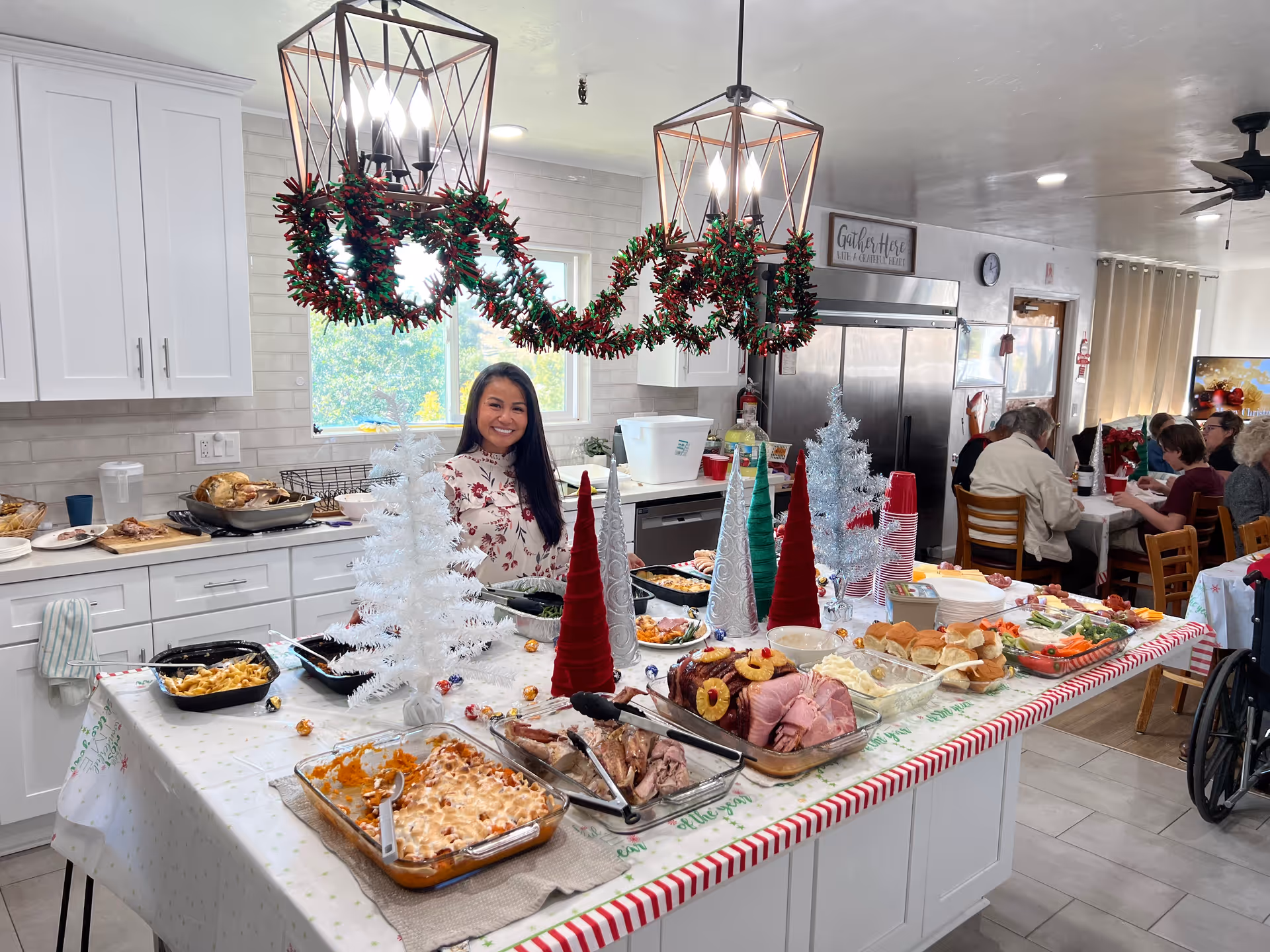 A festive holiday buffet setup in a bright kitchen with white cabinets and modern light fixtures decorated with garlands. A woman stands behind the buffet table smiling. The table is covered with a holiday-themed tablecloth and filled with various dishes including ham, sandwiches, pasta, and vegetables. In the background, several elderly people are seated at tables enjoying a meal in a communal dining area.