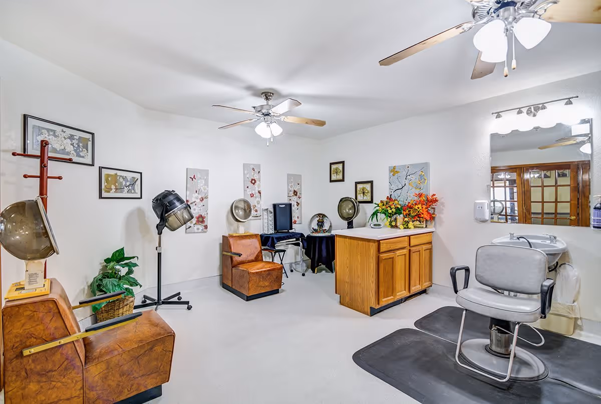 Interior view of a salon area in a senior living facility with two vintage hair dryer chairs, a styling chair in front of a sink and mirror, wooden cabinetry with a countertop decorated with flowers, ceiling fans with lights, framed floral artwork on the walls, and a potted plant in the corner.
