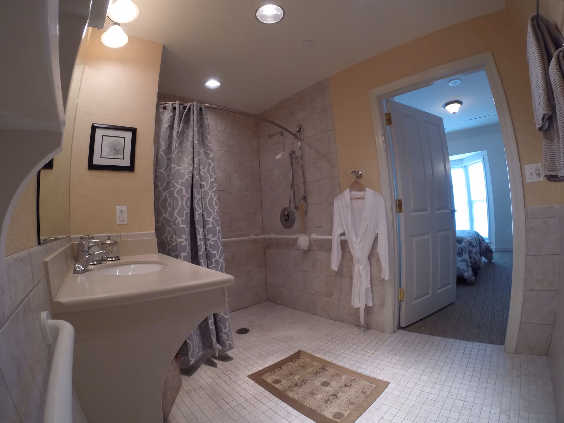 A bathroom with beige tiled walls and floor, featuring a walk-in shower with a gray patterned shower curtain, a white sink with a mirror above it, a white bathrobe hanging on the wall, and an open door leading to a bedroom with a bed and large windows.