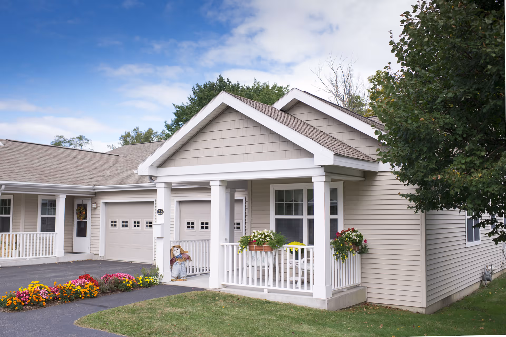 Front view of a single-story senior living apartment building with a covered porch, white columns, beige siding, and colorful flowers.