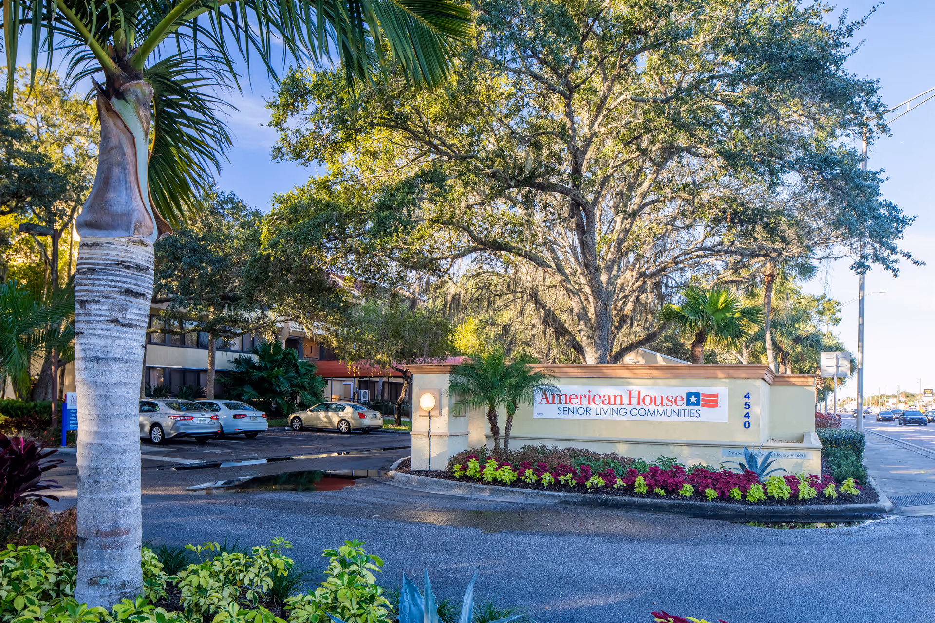 Entrance sign for American House Senior Living Communities surrounded by landscaped plants and trees, with a parking lot and building visible in the background under a clear blue sky.