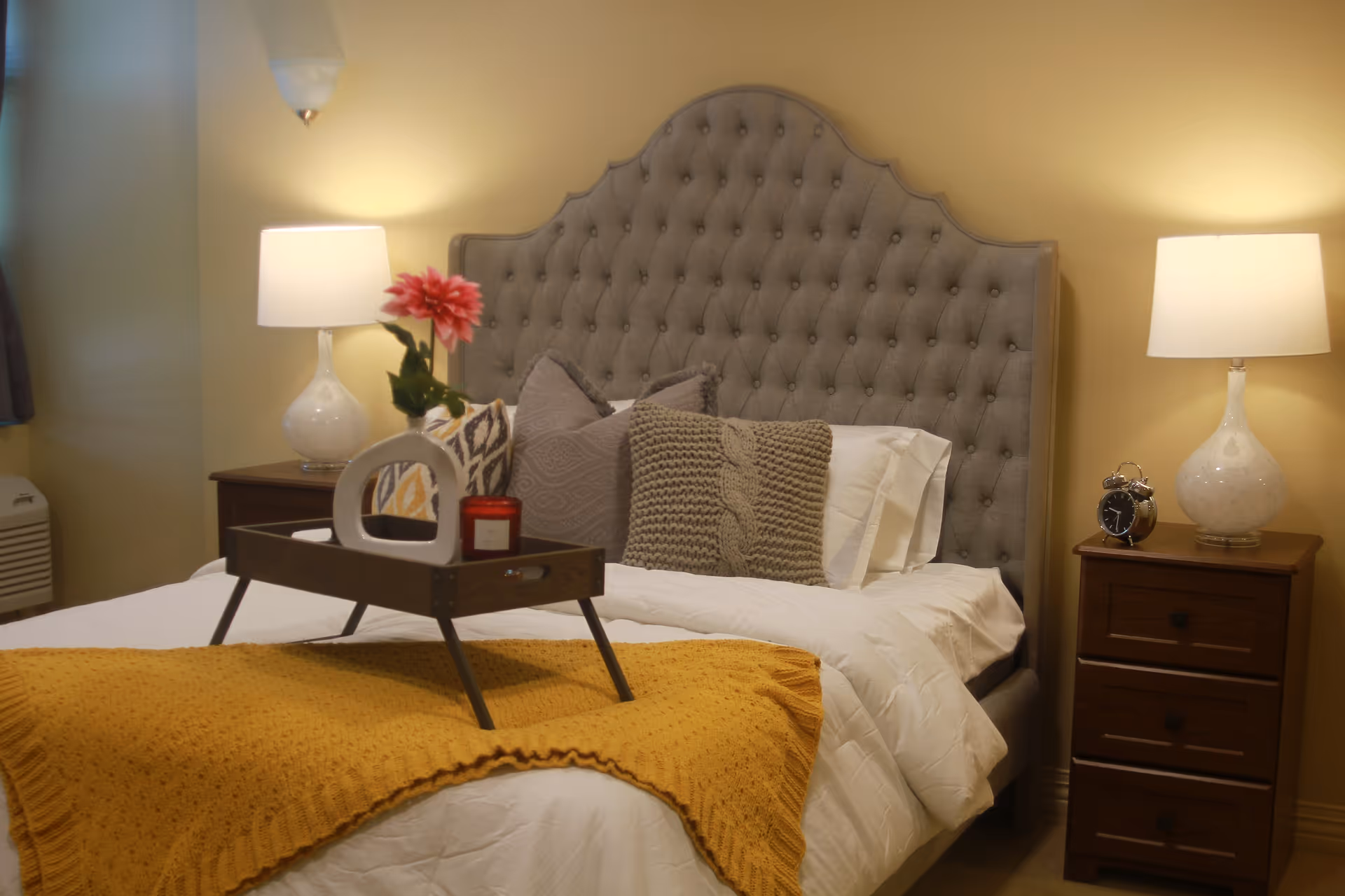 A cozy bedroom with a tufted gray headboard, white bedding, and a mustard yellow knitted throw blanket. There are decorative pillows on the bed and a wooden tray holding a red candle, a flower in a vase, and a decorative object. Two bedside tables each have a white lamp, and one has a small alarm clock.