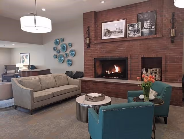 A cozy living room with a beige sofa, two teal armchairs, a round coffee table, and a brick fireplace with a fire burning. Above the fireplace are framed black and white pictures and decorative wall sconces. There are decorative blue plates on the wall and a vase with orange tulips on a small side table.