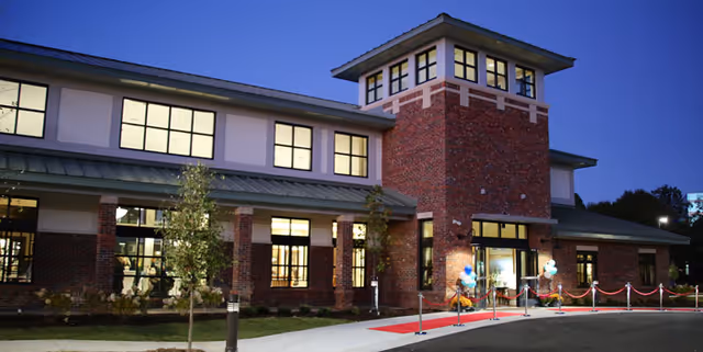 Exterior view of a two-story brick building with large windows illuminated from inside during dusk. The entrance features a red carpet and balloon decorations, with a paved driveway and small trees in front.
