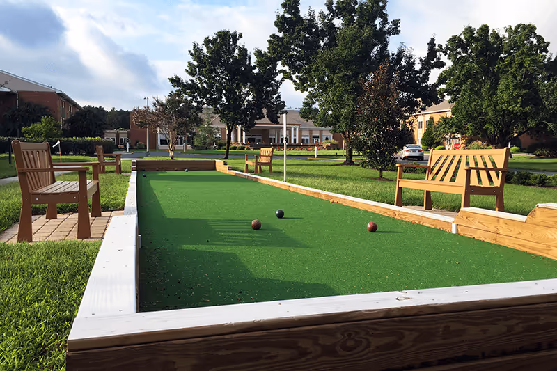 Bocce ball court with green turf, wooden benches, and a brick building in the background.