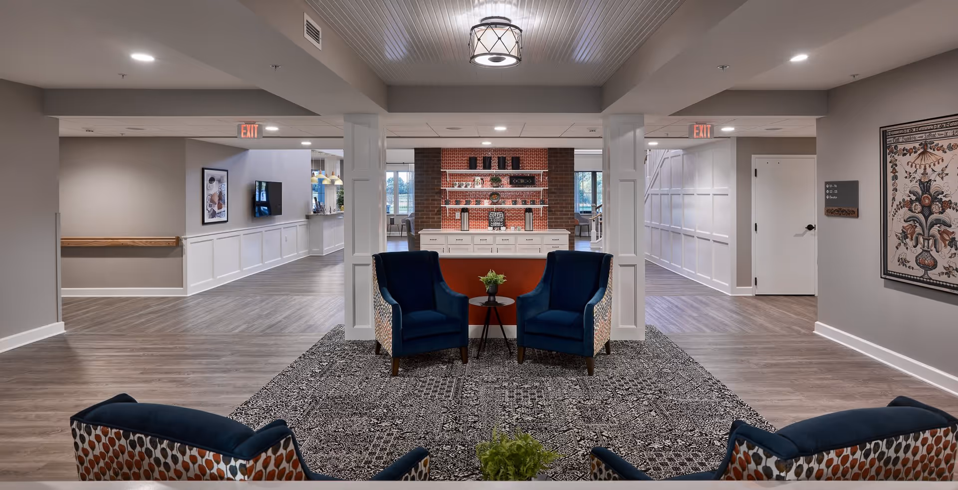 A spacious and well-lit senior living facility common area with four blue upholstered armchairs arranged around a small round table with a plant. The floor has a patterned rug, and the walls are painted light gray. In the background, there is a coffee station with shelves holding cups and decor, and large windows letting in natural light. The ceiling has modern light fixtures, and there are exit signs above doorways on either side.