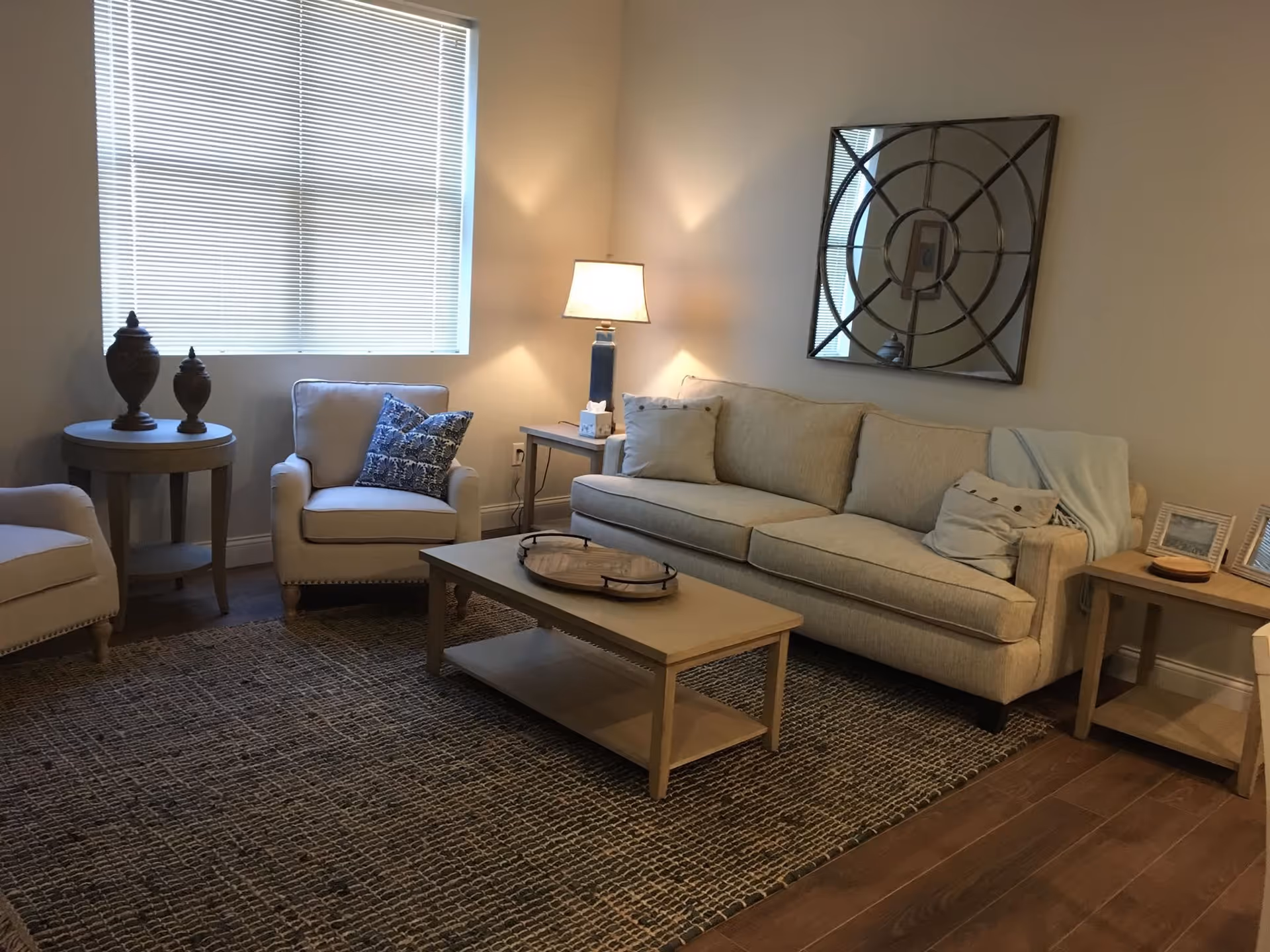A cozy living room with a beige sofa adorned with pillows and a throw blanket, a wooden coffee table with a decorative tray, two beige armchairs, a round side table with two decorative urns, a side table with a lamp and tissue box, a large window with blinds, and a decorative circular mirror on the wall.