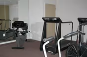 A small fitness room with exercise equipment including a recumbent bike, a treadmill, and a stationary bike. The room has a large mirror on one wall and light-colored walls with carpeted flooring.