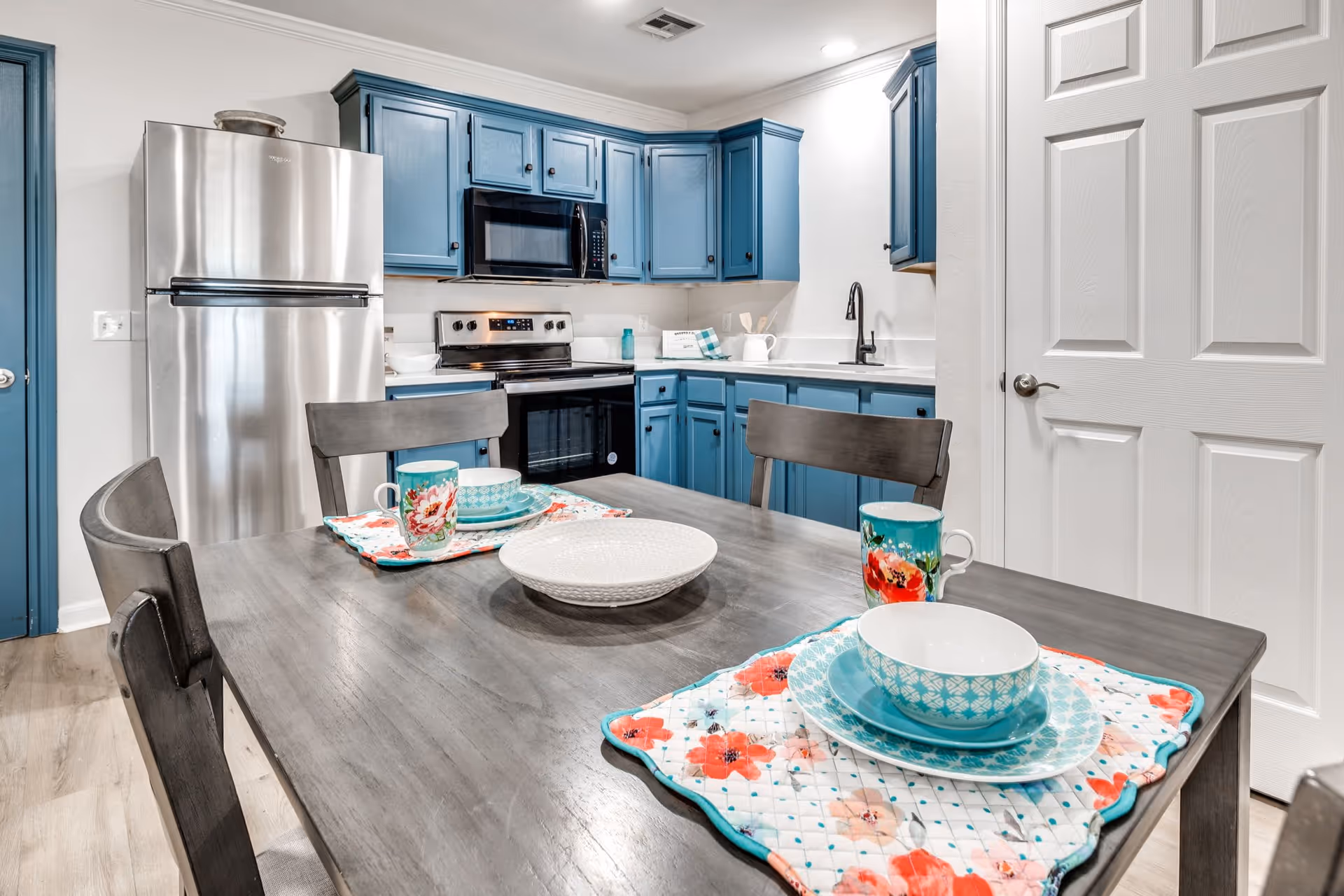 A modern kitchen with blue cabinets, stainless steel refrigerator, stove, and microwave. A wooden dining table with four chairs is set with floral placemats, bowls, plates, and mugs. The kitchen has white countertops and a white door on the right side.