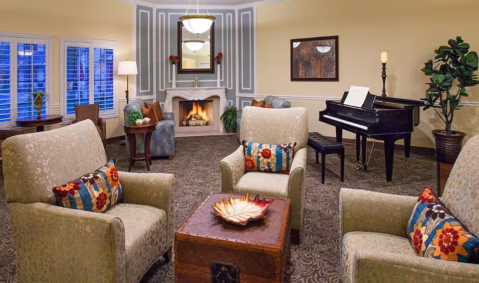 A cozy living room with three upholstered armchairs featuring colorful floral pillows arranged around a wooden chest coffee table. In the background, there is a lit fireplace with a mirror above it, a grand piano with sheet music, a tall floor lamp, and a large potted plant. The room has patterned carpet and light-colored walls with decorative molding.