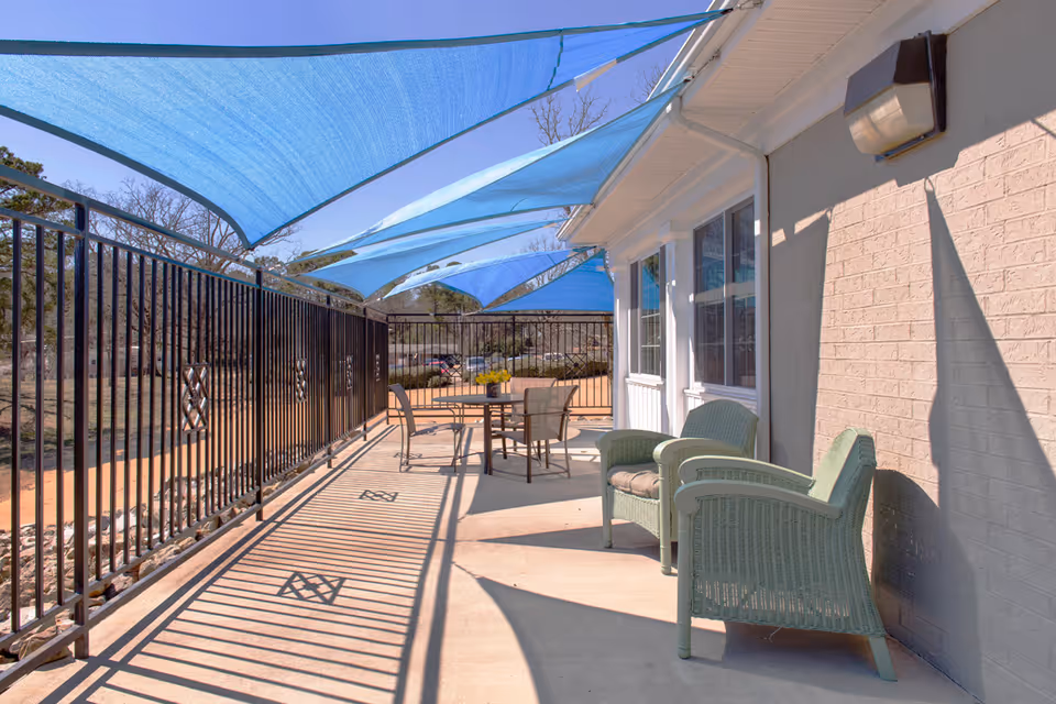 Outdoor patio area with light green wicker chairs and a round table with chairs under blue shade sails attached to the building. The patio is enclosed by a black metal fence and is adjacent to a beige brick building with windows.