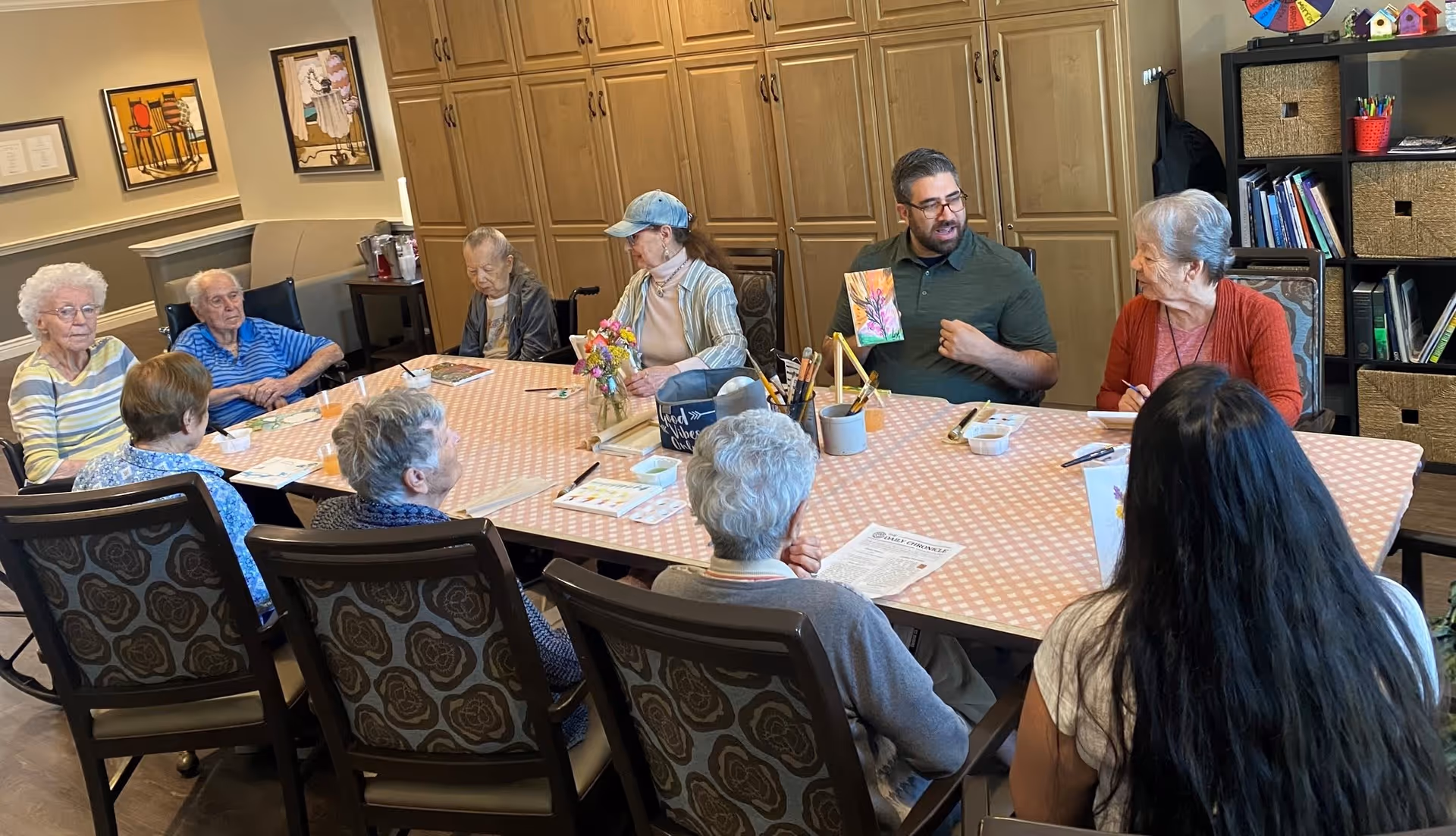 A group of elderly people and a younger man sitting around a large table covered with a pink and white checkered tablecloth in a room with wooden cabinets and shelves. The man is holding up a colorful drawing and appears to be leading an activity or discussion. Various art supplies and papers are on the table.