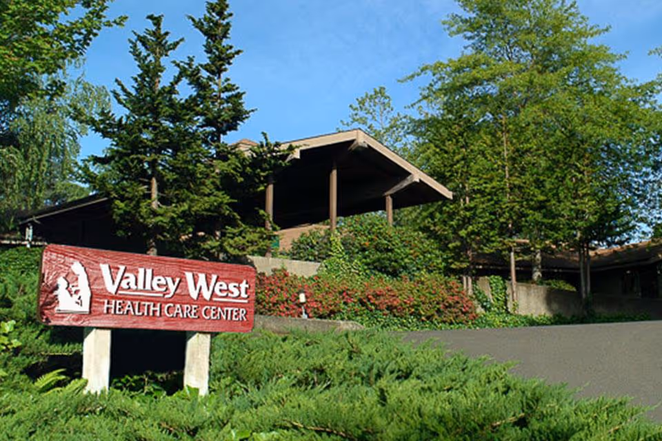 Entrance to Valley West Health Care Center with a wooden sign in front of a low building surrounded by trees and shrubs.