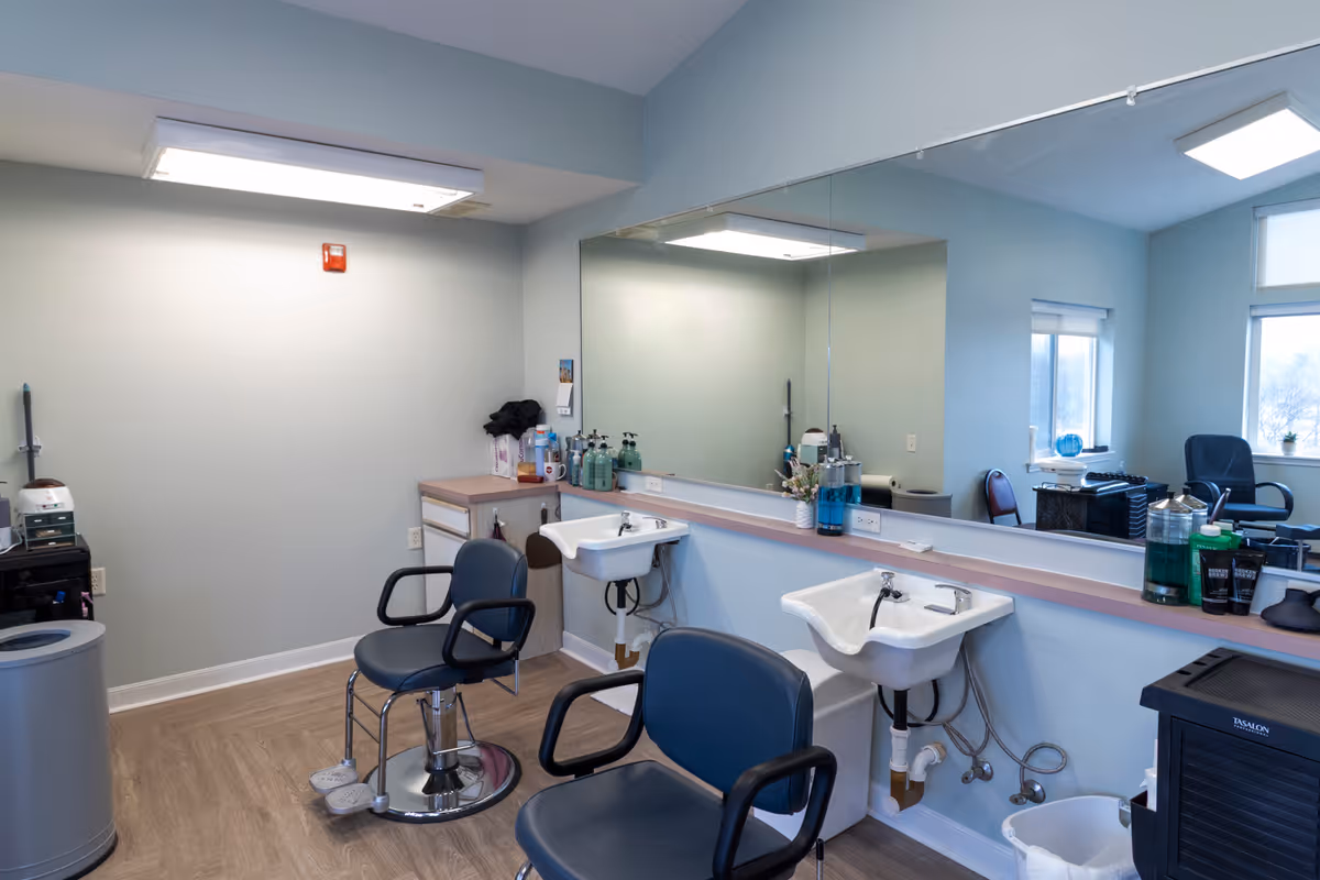 Interior of a salon area in a senior living facility with two black salon chairs in front of two white sinks mounted on the wall. A large mirror spans the wall behind the sinks, reflecting the room. Various hair care products are placed on the counter and a small cabinet is in the corner. The room has light-colored walls and wood flooring.