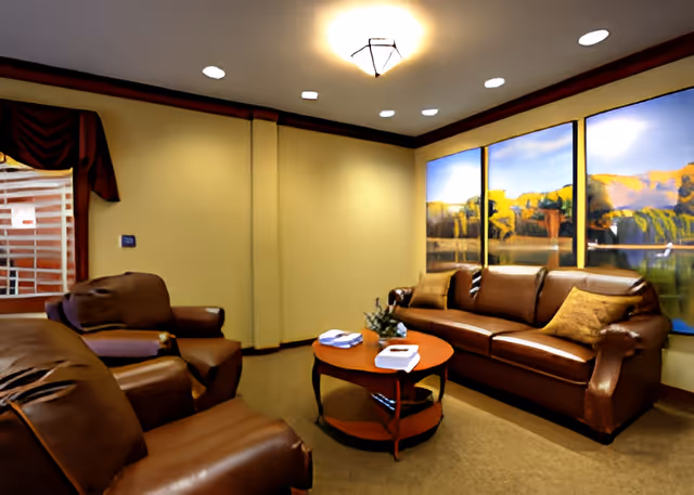 A cozy living room with brown leather sofas and armchairs arranged around a round wooden coffee table. The walls are painted beige with a large window featuring a scenic landscape view. The room is softly lit with ceiling lights.