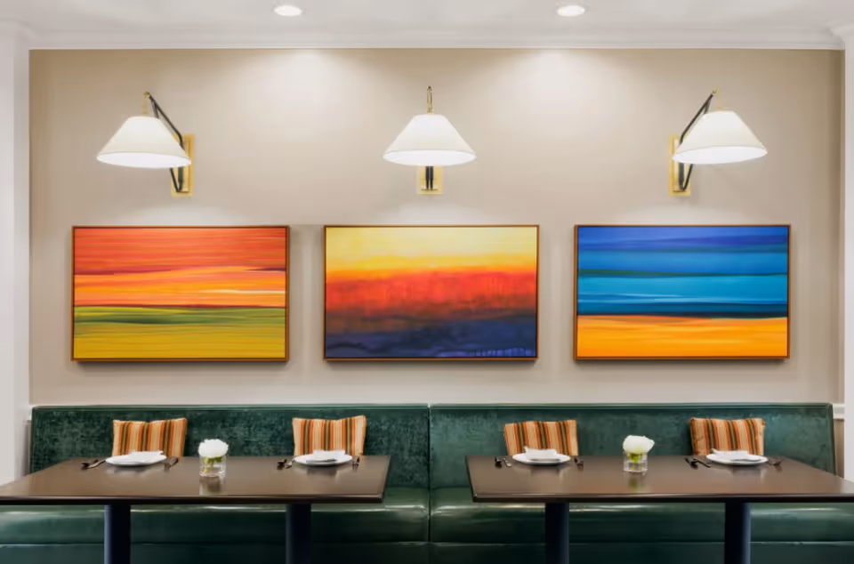 A dining area with two dark wooden tables set with white plates, silverware, and small white flower centerpieces. The seating consists of a green upholstered bench with striped orange and beige pillows. Above the bench are three colorful abstract paintings in rectangular frames, illuminated by three wall-mounted lamps with white shades.