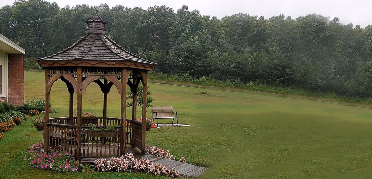 A wooden gazebo with a shingled roof surrounded by pink flowers on a grassy lawn. There is a wooden bench on a small concrete pad in the background, with a line of trees further behind. Part of a brick building is visible on the left side.