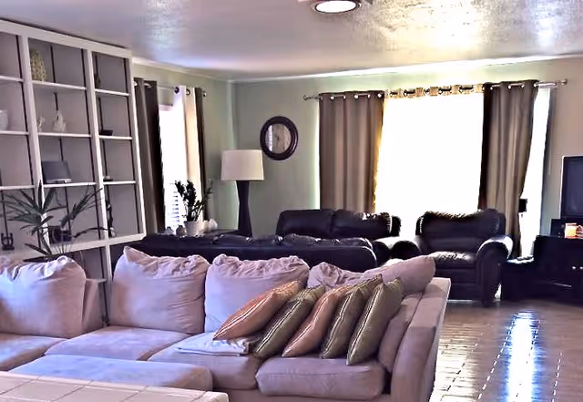 A spacious living room with a large beige sectional sofa adorned with multiple pillows in the foreground. Behind it, there are two dark leather armchairs and a matching sofa. The room features a tall floor lamp, a round wall clock, and large windows with beige curtains allowing natural light to enter. Built-in shelves with decorative items and plants are visible on the left side, and a television is placed on a stand to the right.
