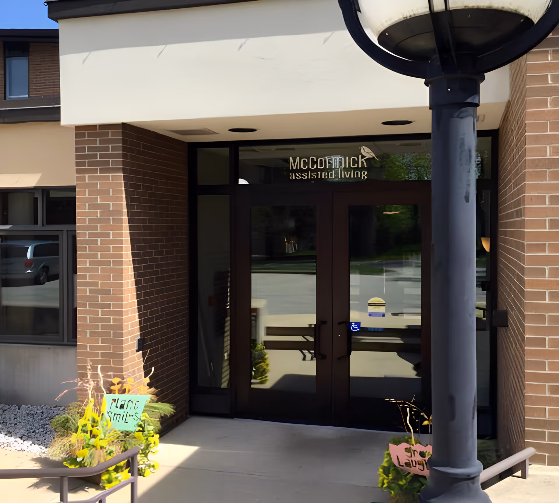 Entrance to McCormick Assisted Living facility with double glass doors, brick walls, a lamp post, and small plants with signs near the entrance.